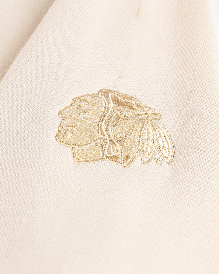 beige cream TravisMathew hoodie with tiny wordmark logo across chest and Chicago Blackhawks primary logo embroidered on left sleeve - logo lay flat